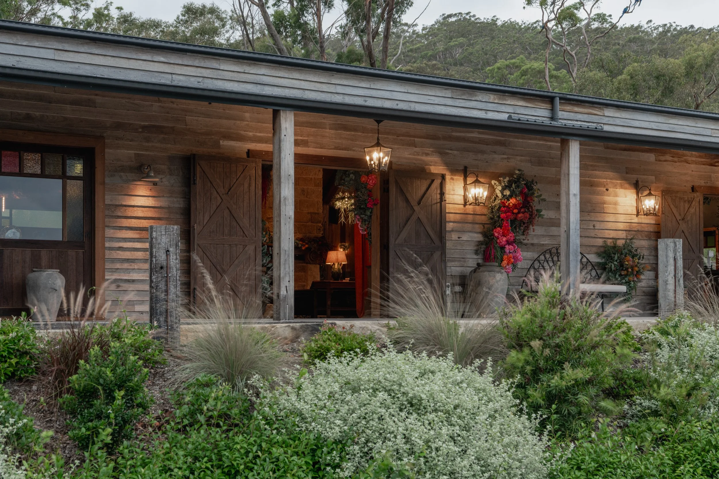 Front view of The Barn at Saddles Mount White with open double doors, surrounded by greenery and plants, with three lantern-style lights illuminating the porch area.