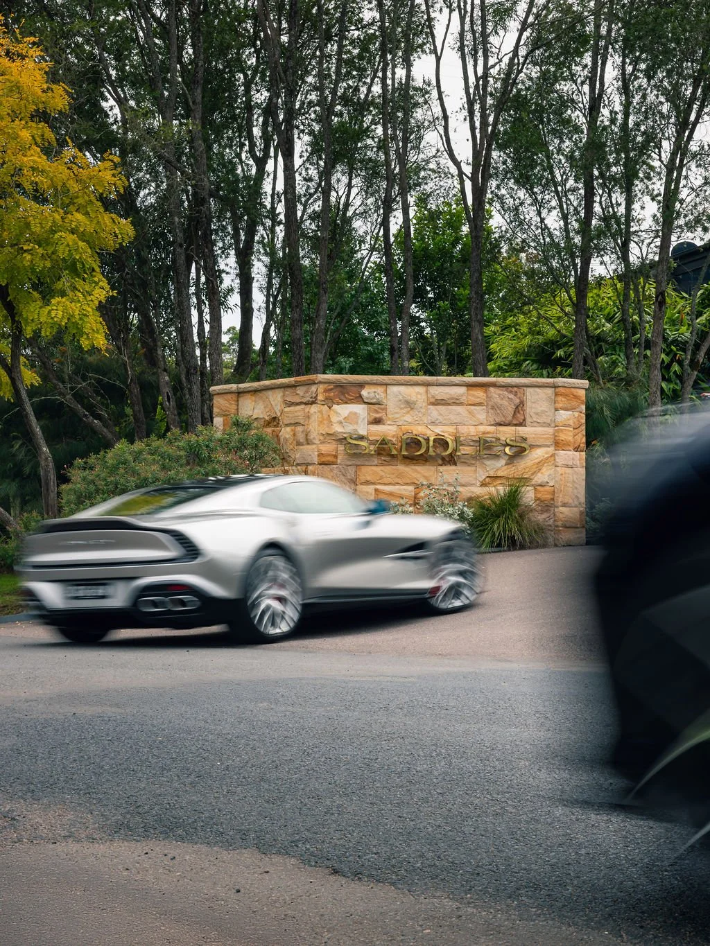 A silver sports car driving past a stone entrance sign that reads 'SADDLE' with trees and greenery in the background.