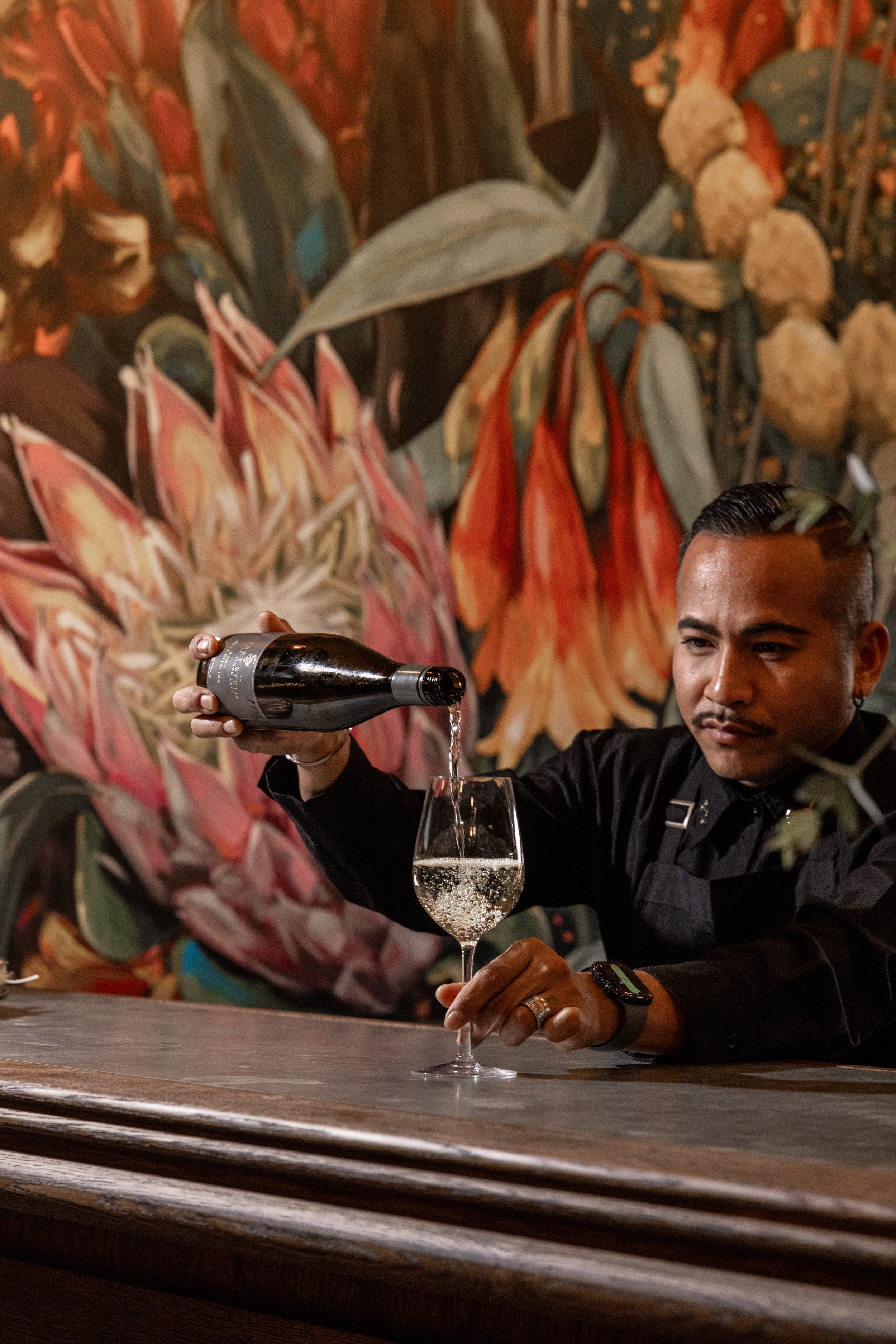 A bartender at The Barn at Saddles Mount White pouring a glass of sparkling wine at a bar, with a colorful floral painting on the wall behind him.