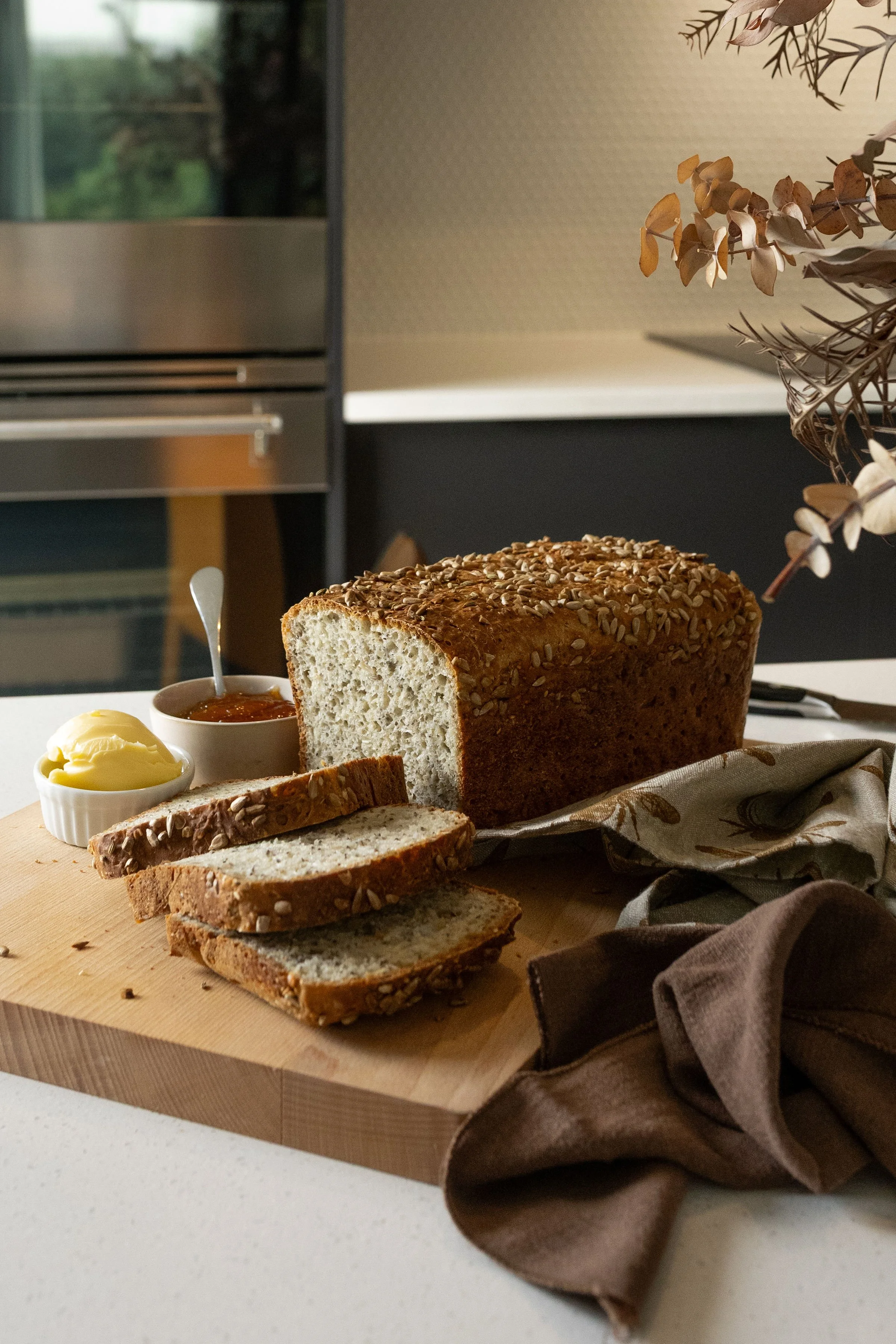 Sliced multigrain bread on a cutting board with butter and jam, in a modern kitchen setting.