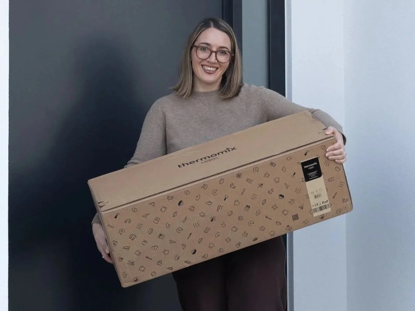 A young woman with glasses, smiling, holding a large cardboard box labeled 'thermomix' and decorated with small icons, standing indoors near a dark wall.