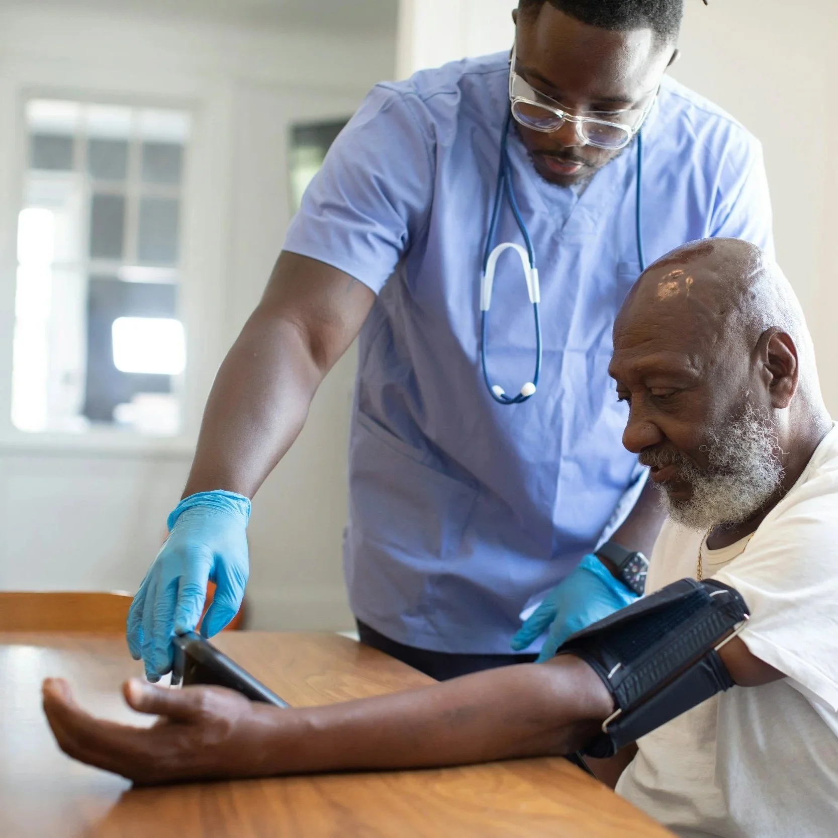 A healthcare professional, wearing scrubs, glasses, and a stethoscope, provides medical assistance to a patient who is sitting at a table, holding a smartphone, with a blood pressure cuff on his arm.