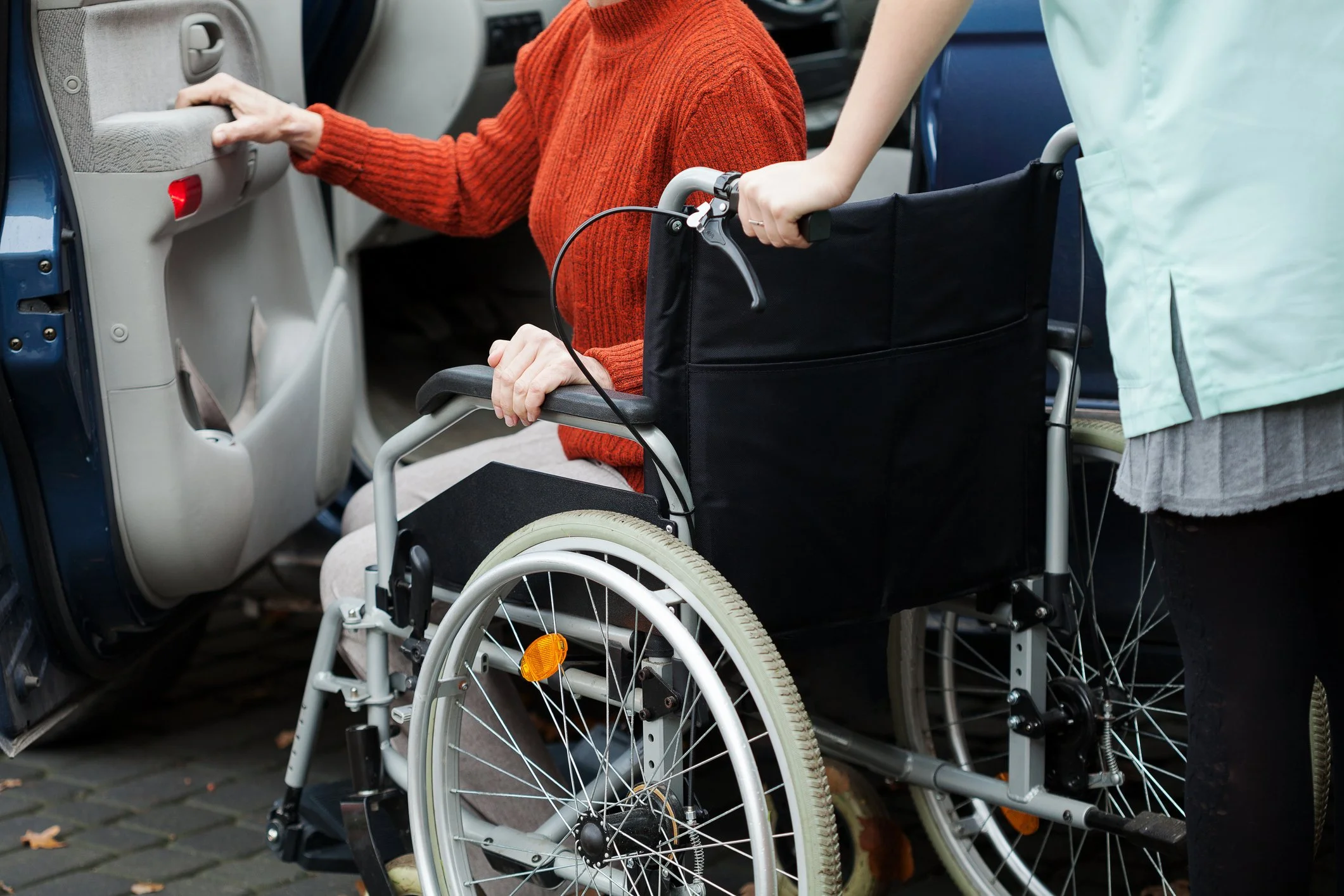 A person in a wheelchair is being pushed towards a vehicle by a caregiver or nurse. The person in the wheelchair is wearing a red sweater and is reaching for the vehicle's door handle.