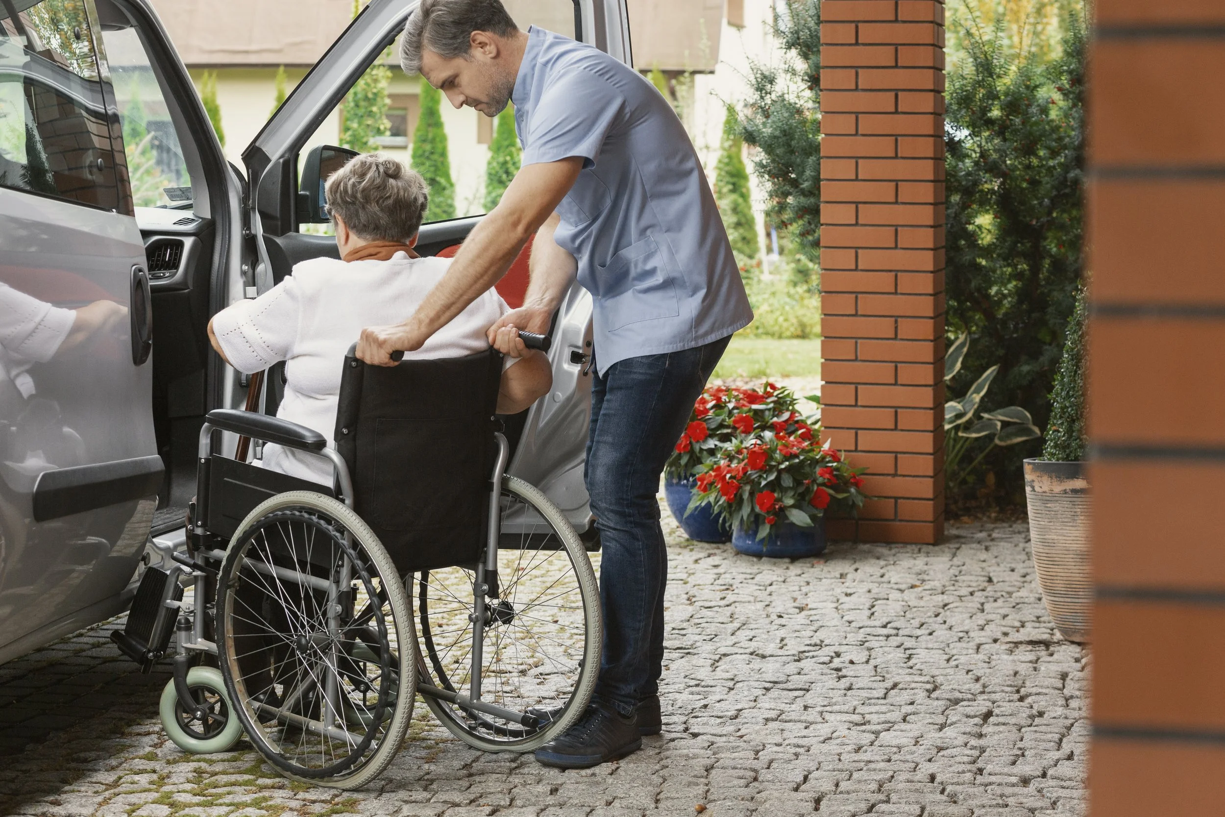 A man in a wheelchair being assisted by a caregiver to get into an SUV parked outside a brick building with potted plants and greenery nearby.