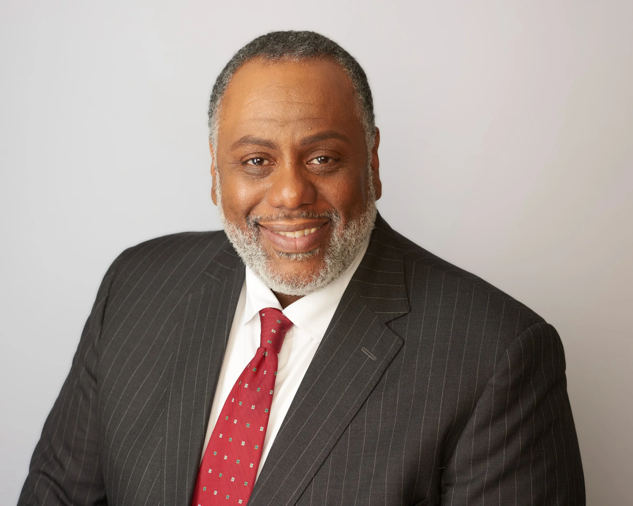 Portrait of a smiling African American man with gray hair and beard, wearing a dark pinstriped suit and red tie, against a plain light background.