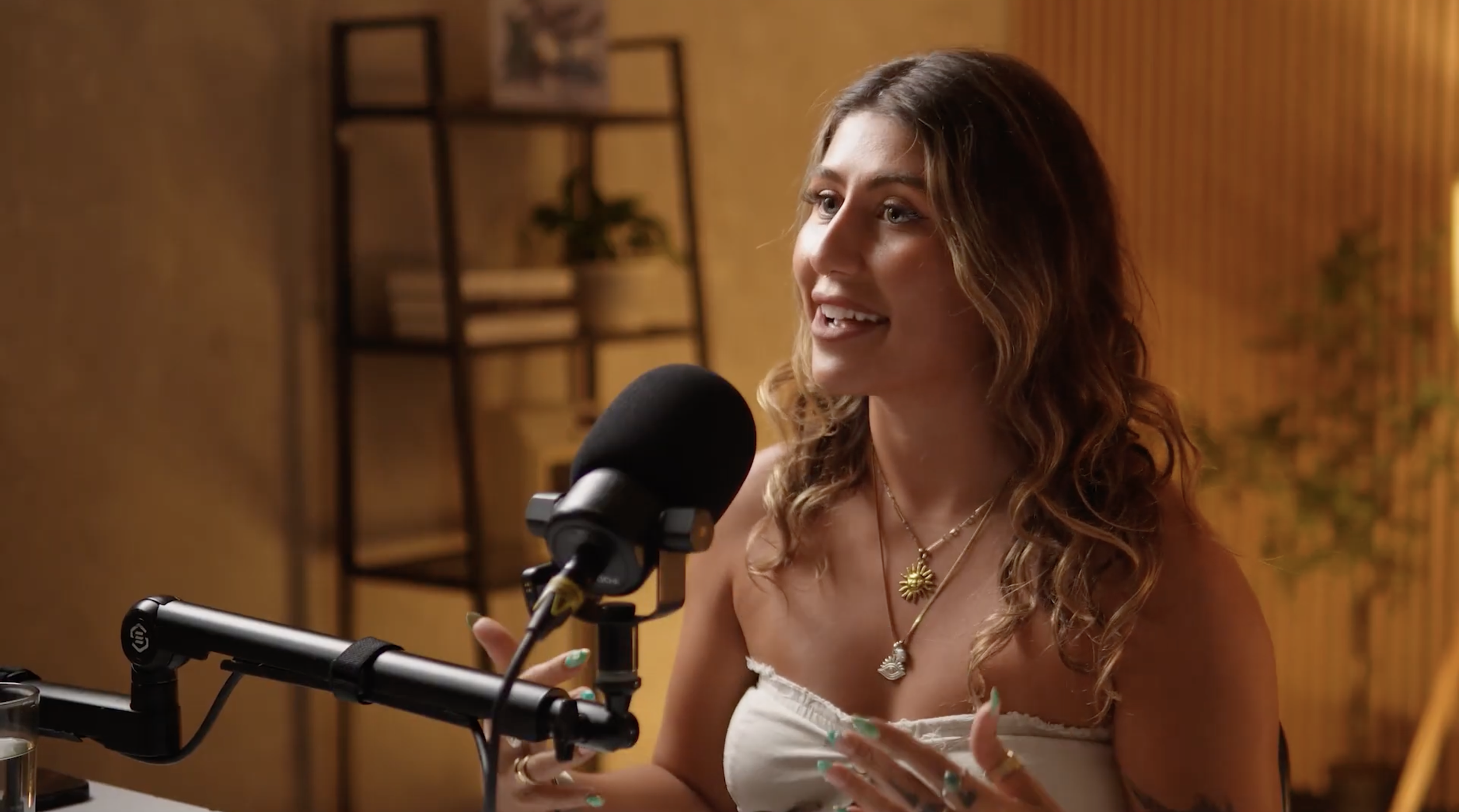 A woman with long curly hair talking into a microphone in a recording studio, wearing a white top and jewelry.
