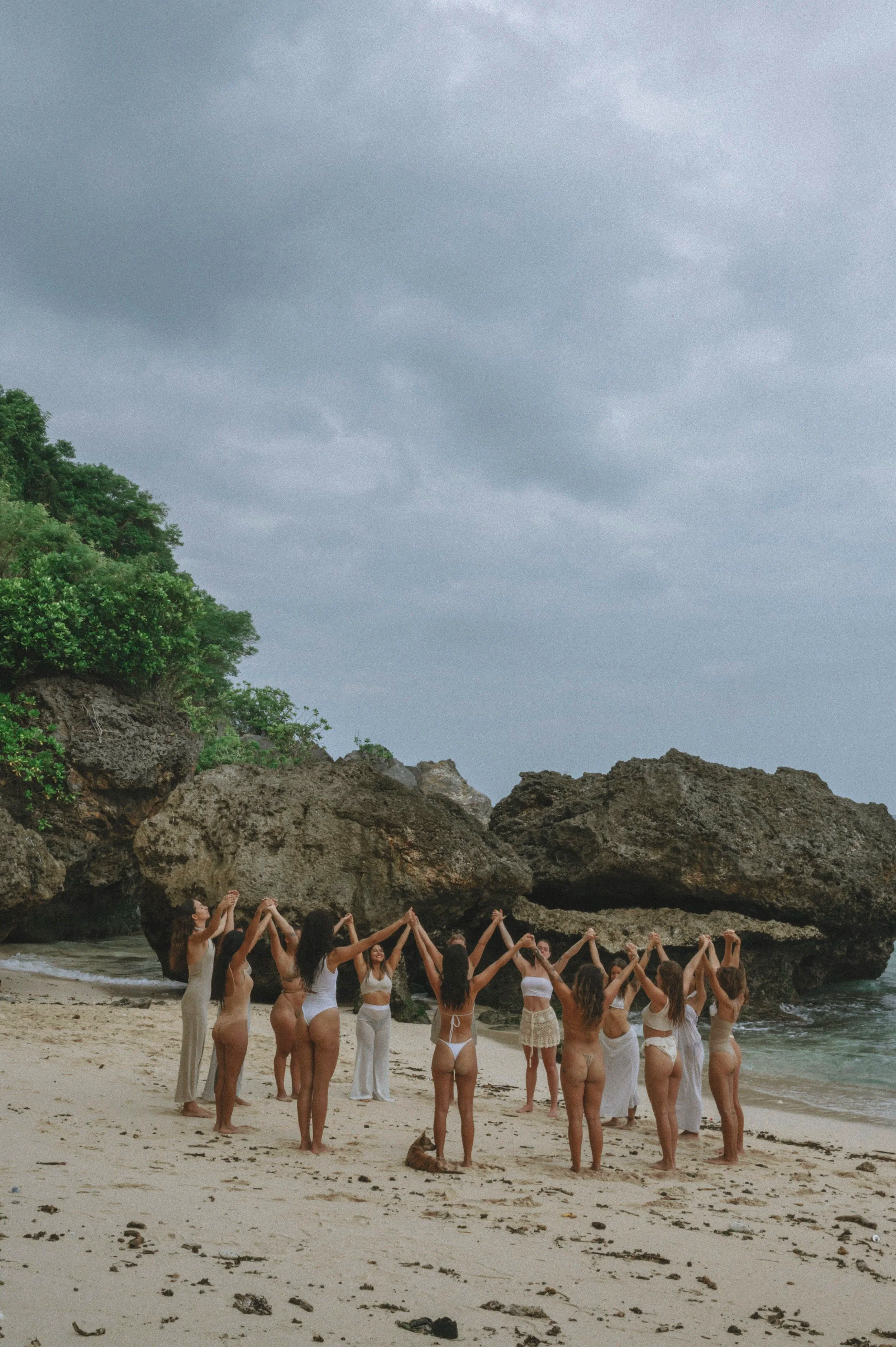 Women dressed in white or beige standing on a beach, holding hands in a circle, facing a large rock formation with trees on top, under an overcast sky.