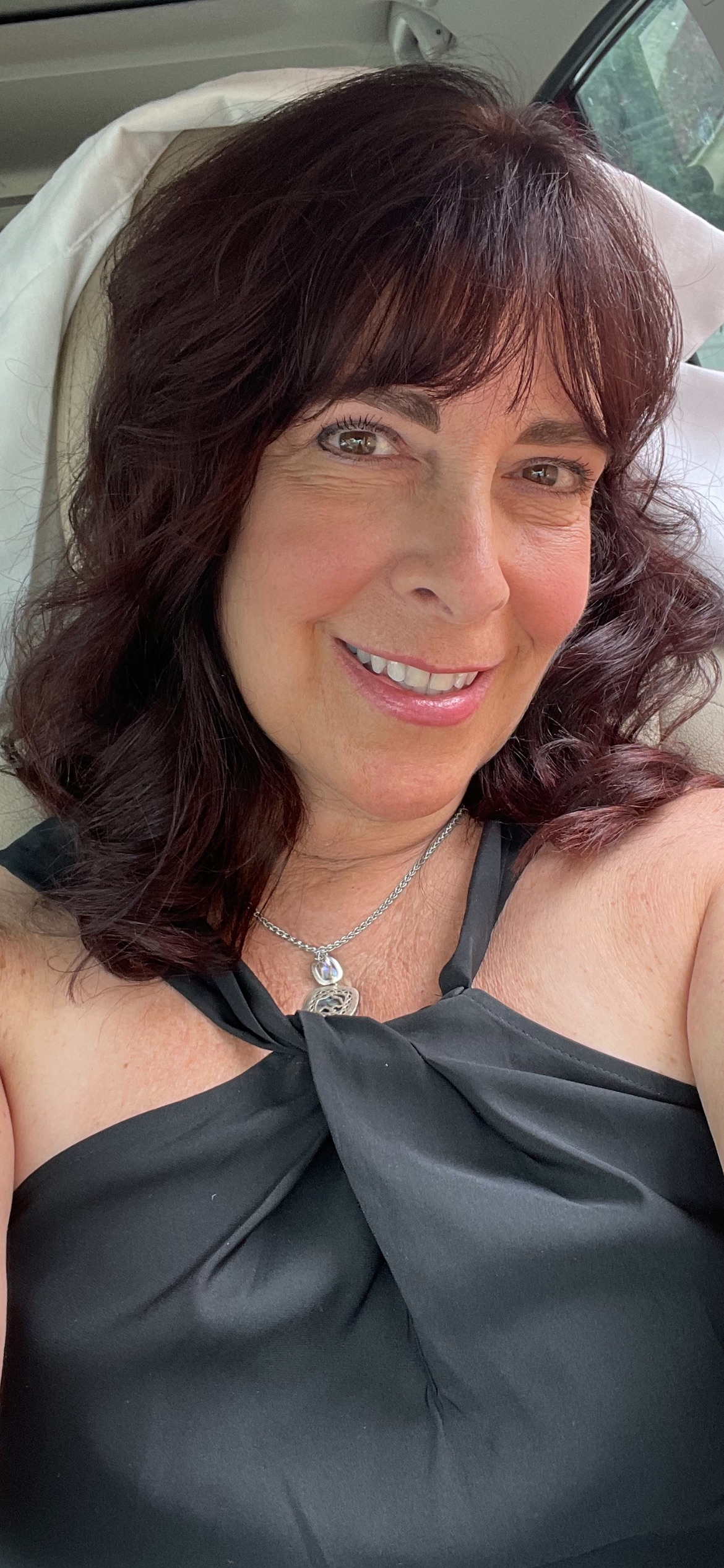A woman with wavy, dark red hair smiling for a selfie, wearing a black dress with a tied neckline and a silver necklace, sitting in a vehicle with a white headrest behind her.
