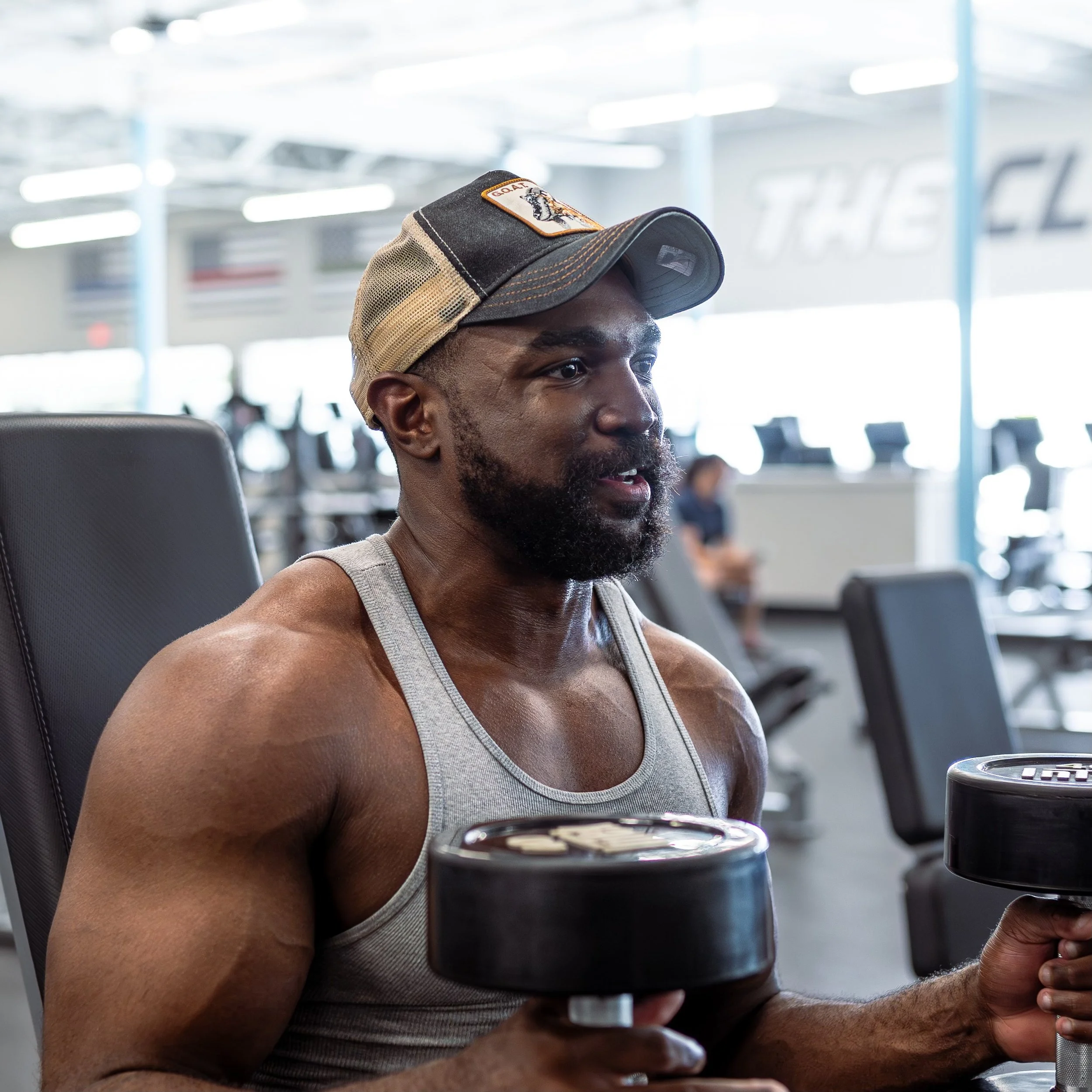 A man with a beard and a sleeveless shirt sitting in an airport or public seating area. He is wearing a baseball cap with a patch that says 'GOLD' and an image of a tiger on it. He is holding a cup or container, and the background shows blurred airport or terminal seating and architecture.