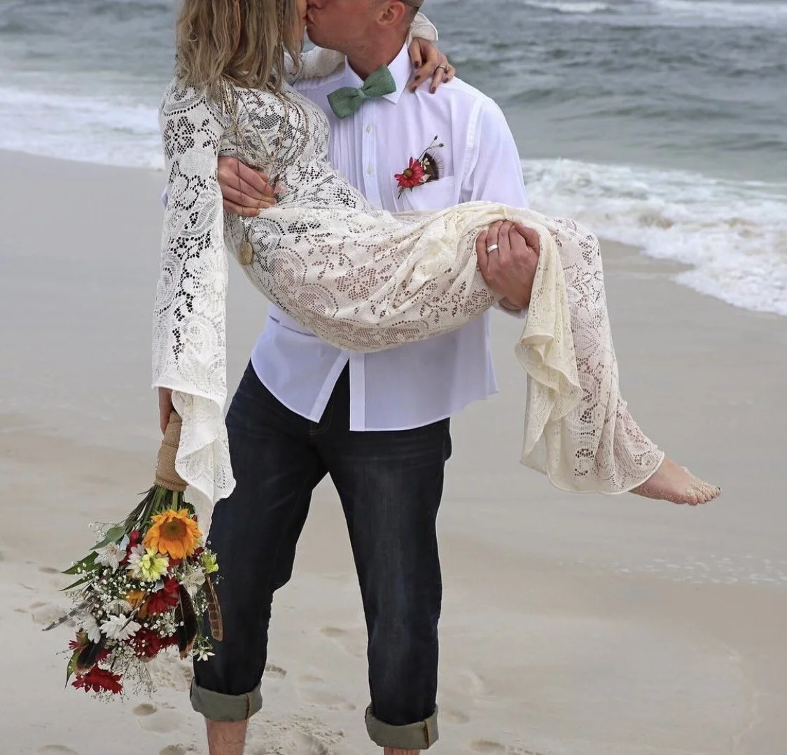 A man in a white shirt and green bowtie holding a woman in a lace dress, while she holds a bouquet of flowers, on a sandy beach with ocean waves in the background. The man is kissing the woman.