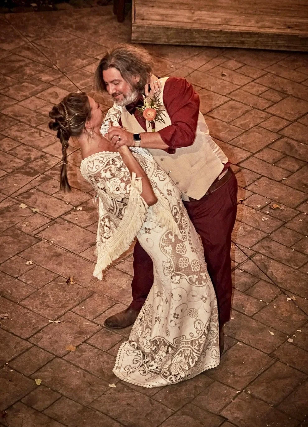 A man and woman dance closely on a brick-paved floor, celebrating a special occasion, likely a wedding. The woman wears a white lace dress and has her hair styled in an updo, while the man wears a beige vest over a maroon shirt with rolled-up sleeves, and has a boutonniere on his vest.