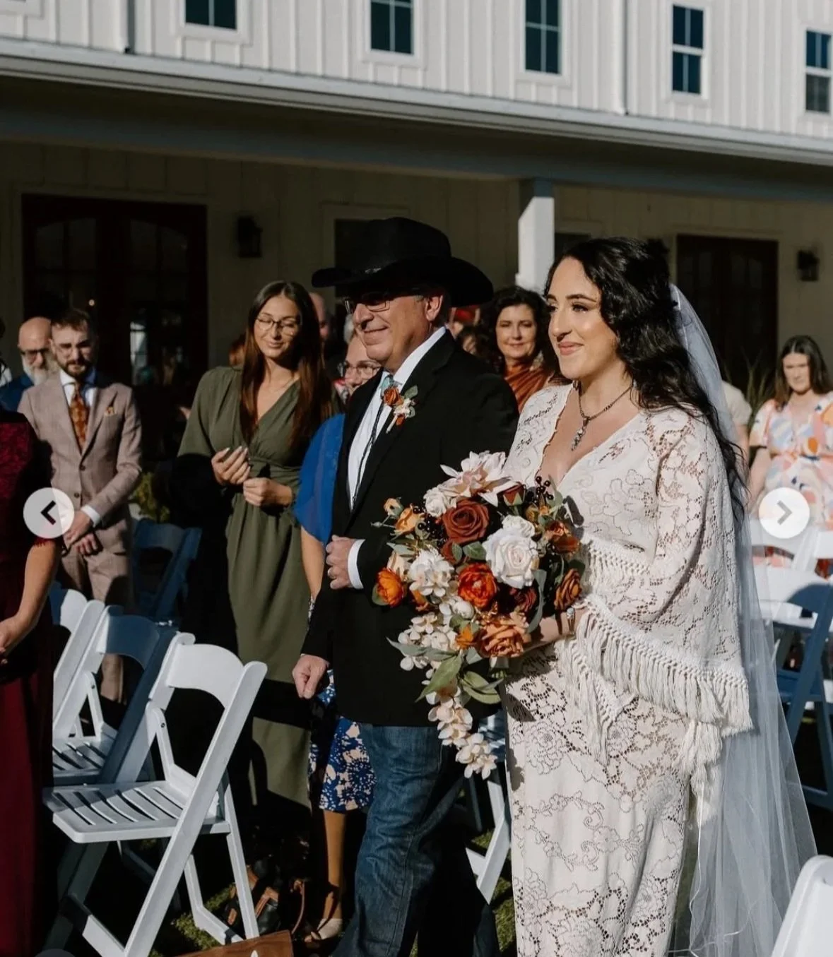 A bride with long dark hair, holding a bouquet of flowers, walking down the aisle with an older man in a black cowboy hat, at an outdoor wedding ceremony with several guests in the background.