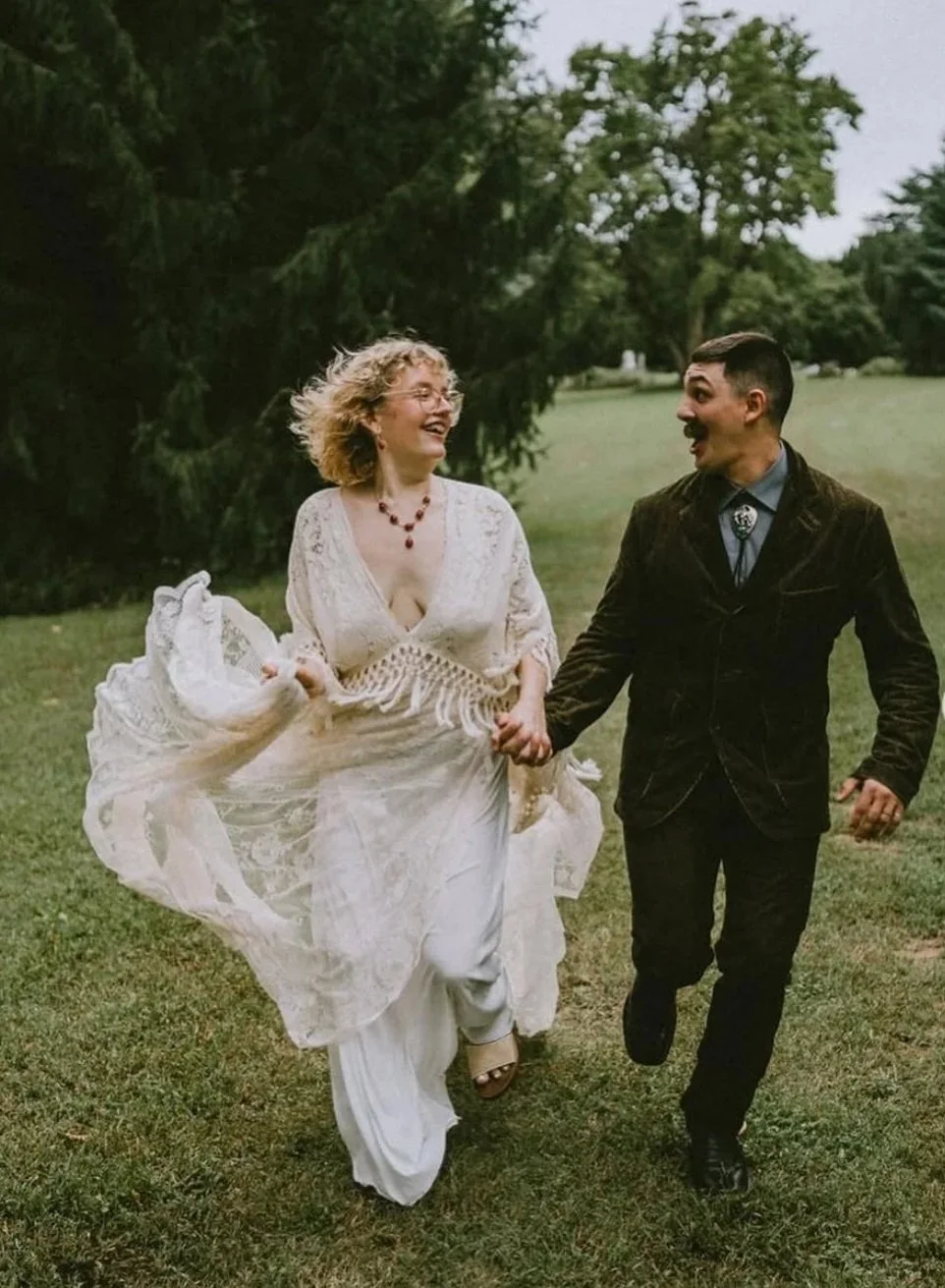 A couple running outdoors, smiling and holding hands. The woman is wearing a white dress and the man is in a dark suit. They are in a grassy park with trees in the background.