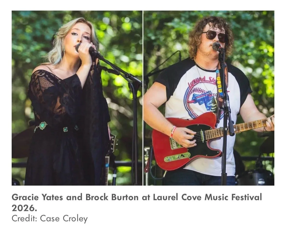 Two musicians performing at an outdoor music festival; a woman singing into a microphone on the left, a man playing an electric guitar and singing into a microphone on the right. Trees in the background.