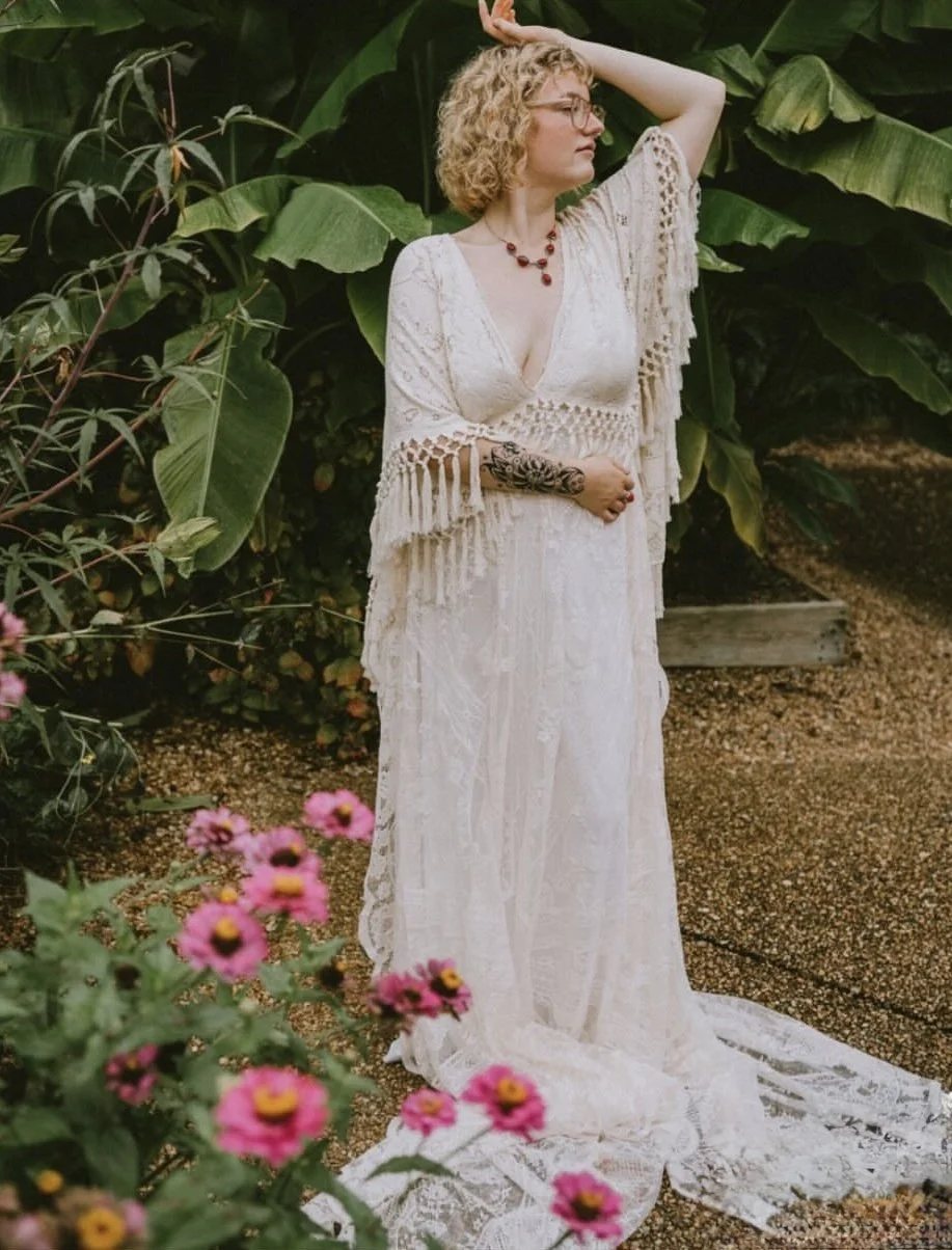 A woman with curly blonde hair and glasses wearing a bohemian-style white lace dress with fringe details, standing outdoors among lush green plants and pink flowers, posed with one arm raised and her gaze to the side.