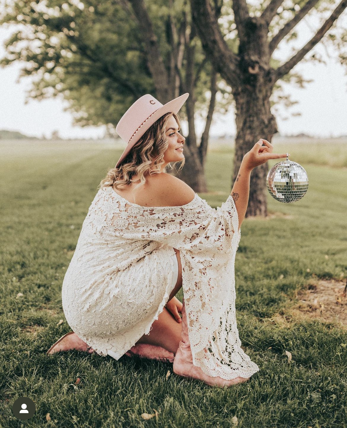 Woman squatting on grass holding a disco ball outside in a park with trees in the background, wearing a wide-brimmed pink hat, a lace dress, and pink boots.