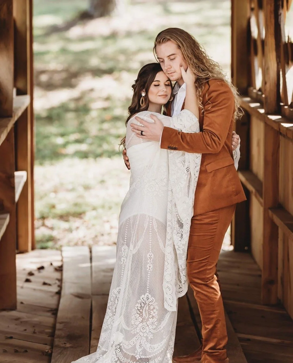A woman in a white lace dress and a man in a brown suit embracing each other in a wooden outdoor setting.