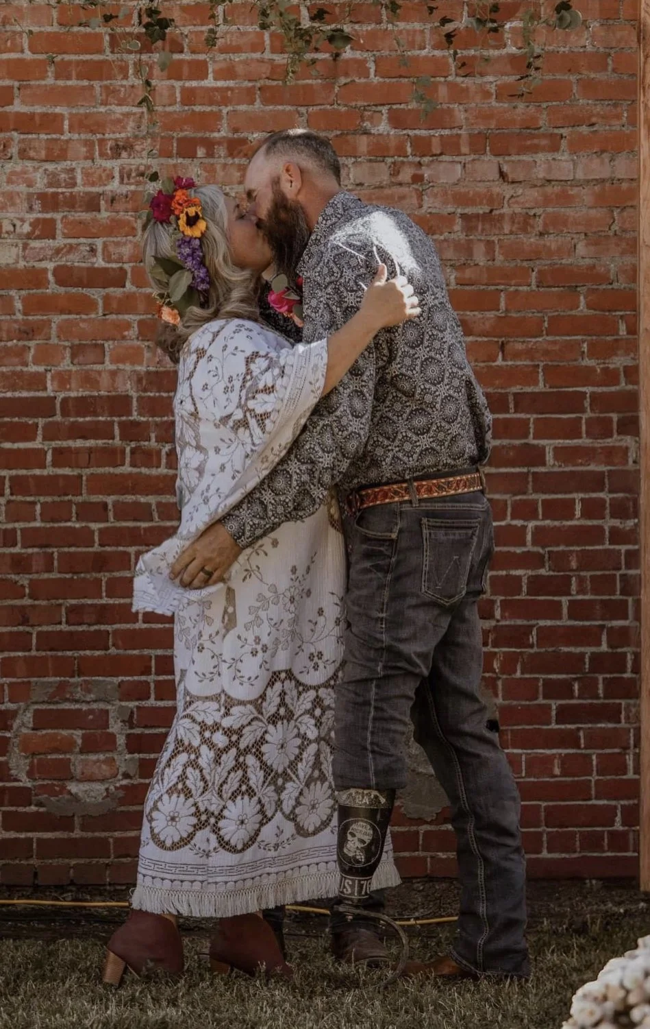 A couple kissing in front of a brick wall. The woman is wearing a white embroidered dress and a floral headband, and the man is dressed in a patterned shirt and jeans.