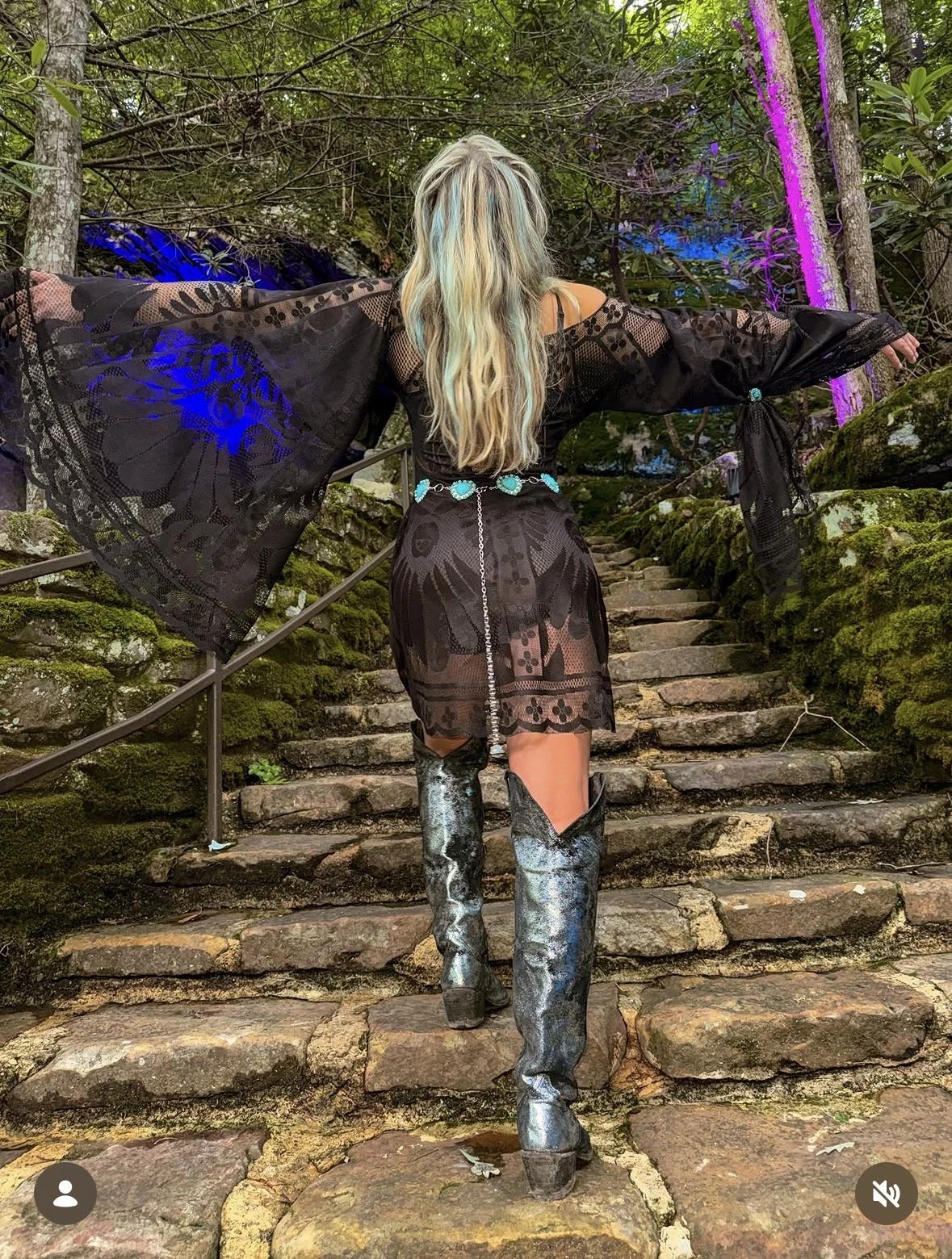 Person with long hair wearing a sheer black dress with lace details, black underwear, a blue belt, and metallic cowboy boots, walking up stone steps in a wooded area with colorful lights.
