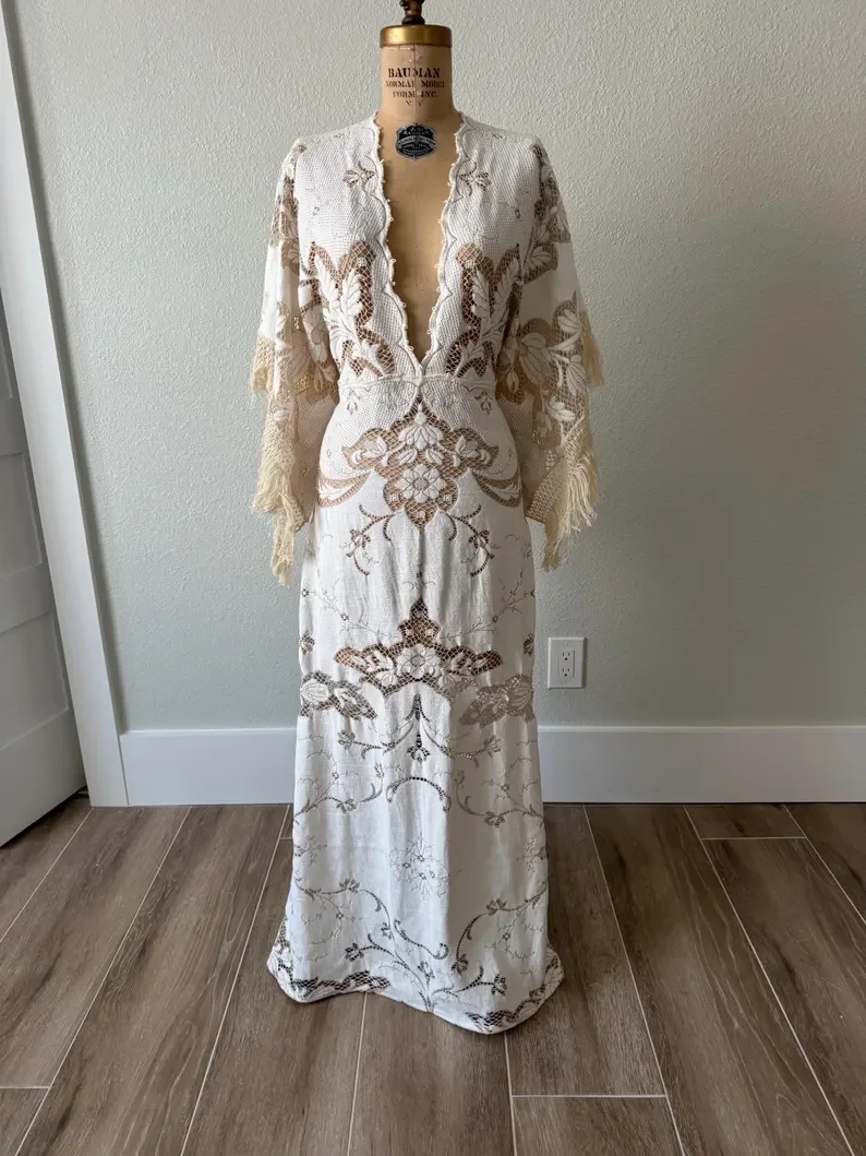 A vintage dress form displaying a long, cream-colored lace dress with intricate floral and geometric embroidery. The dress has a deep V neckline, long sleeves, and a flowing skirt, set against a plain wall with a white electrical outlet visible on the right.
