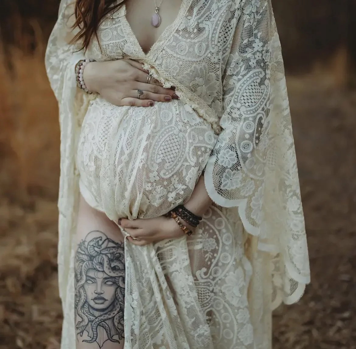 A woman wearing a cream-colored lace dress, holding her pregnant belly with one hand, and revealing a detailed tattoo of Medusa on her thigh. She is accessorized with jewelry, including rings, bracelets, and a necklace with a purple pendant.