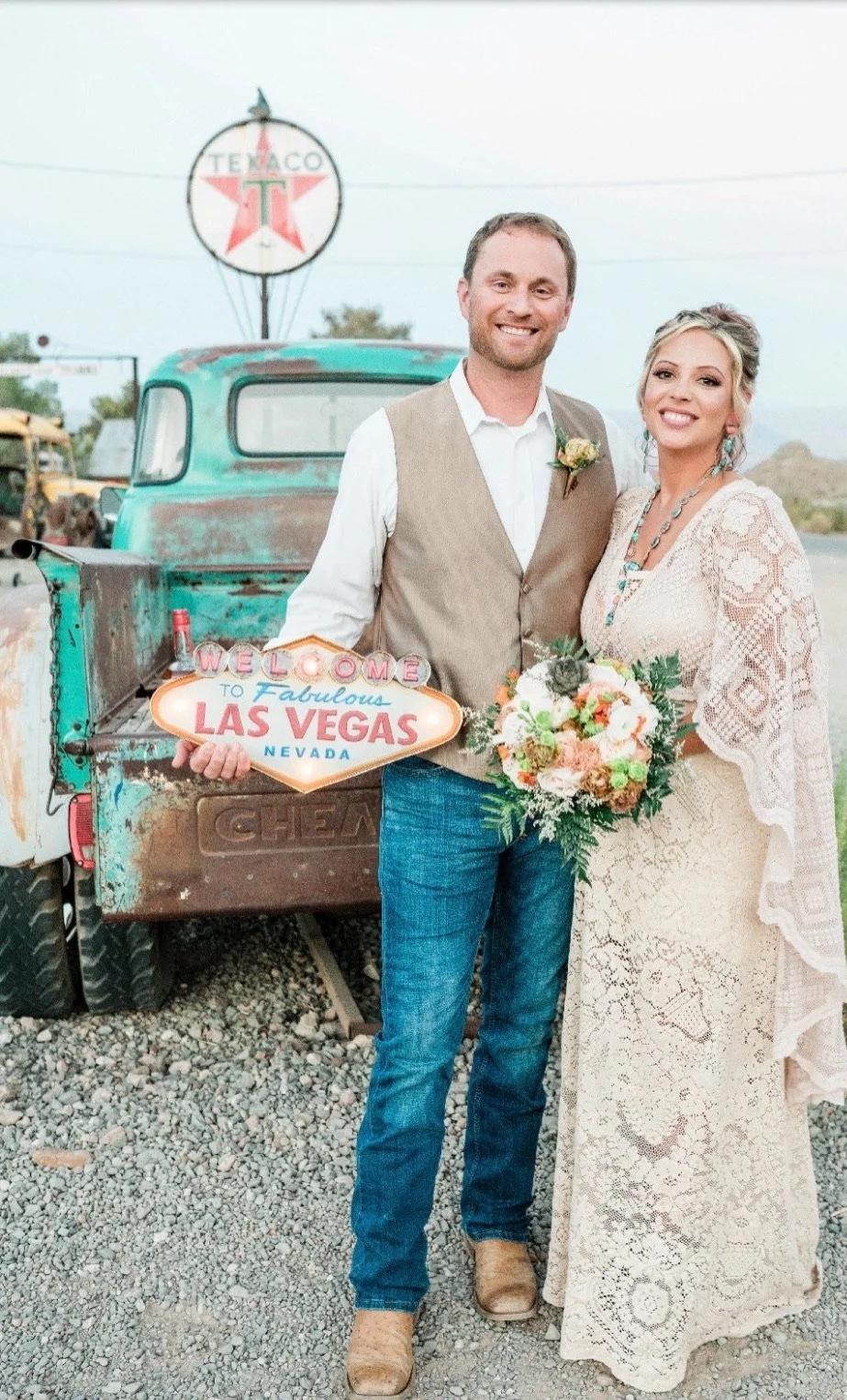 A smiling man and woman in wedding attire standing in front of a vintage truck with a Texaco sign in the background. The man is holding a "Welcome to Fabulous Las Vegas Nevada" sign, and the woman is holding a bouquet of flowers.
