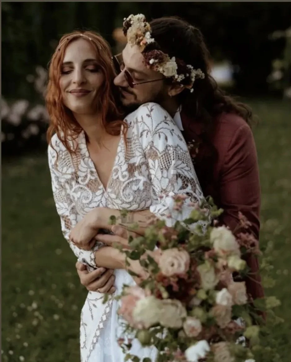 A couple embracing outdoors at sunset, with the man wearing a floral crown and glasses, the woman in a white lace dress holding a large bouquet of pink and white flowers.