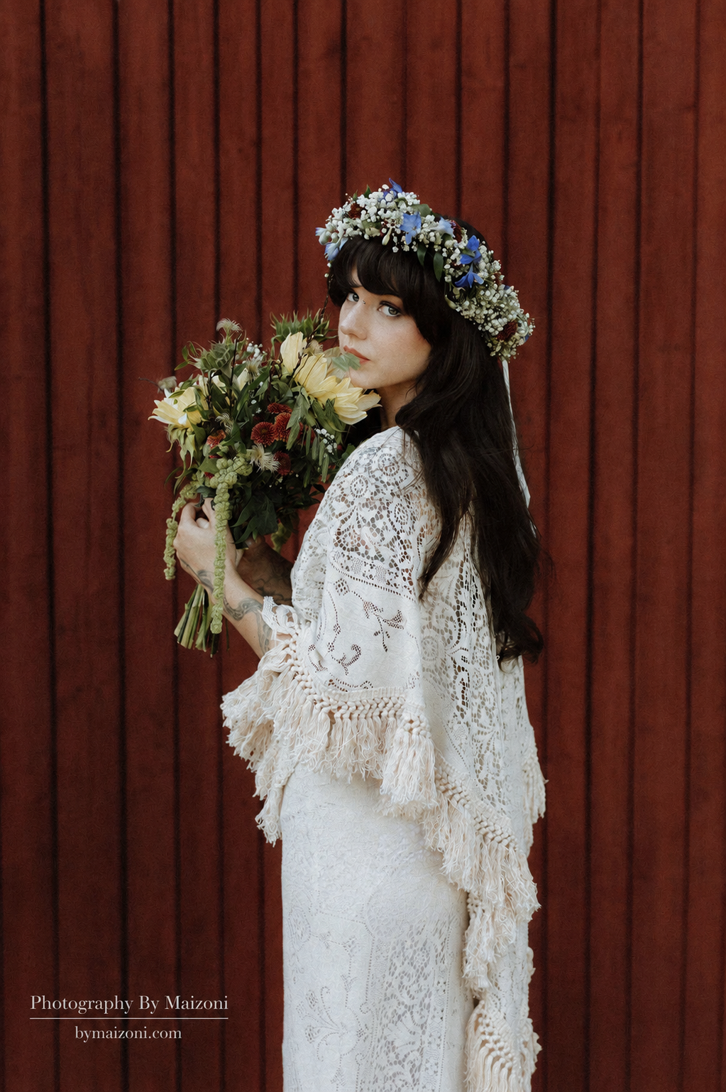 A woman with long black hair wearing a white lace dress and a floral crown, holding a bouquet of flowers, standing in front of a wooden background.