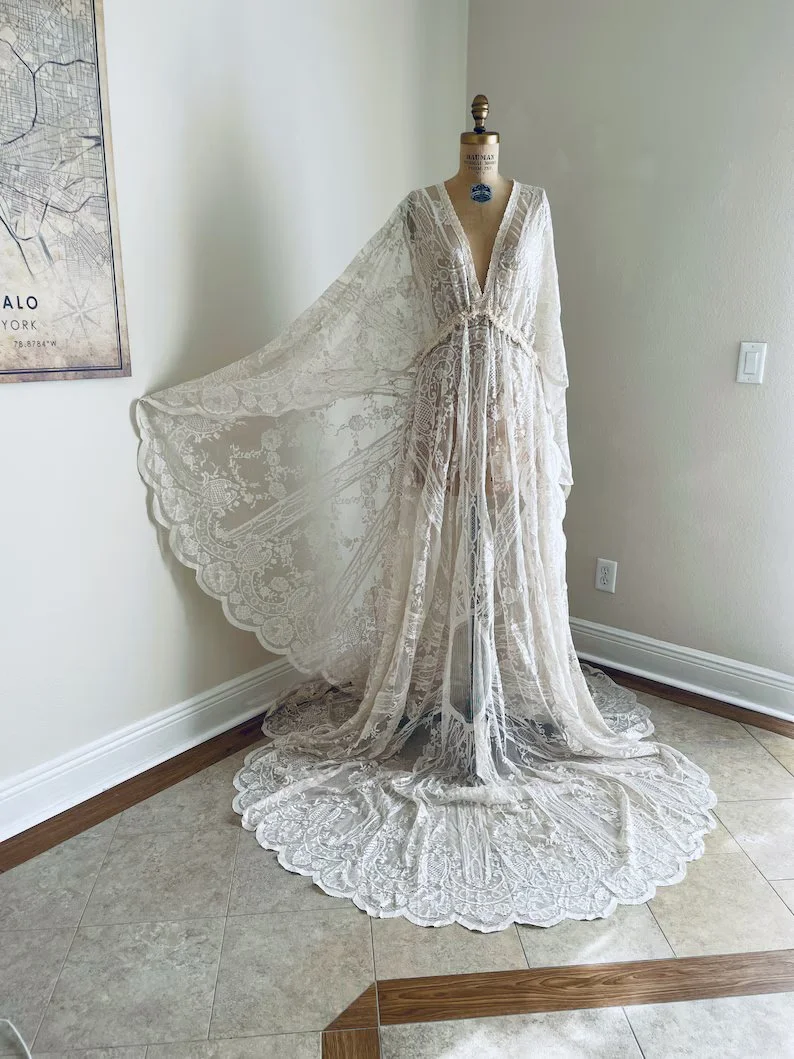 Elegant cream-colored lace wedding dress with a long train and sheer cape displayed on a mannequin in a room.