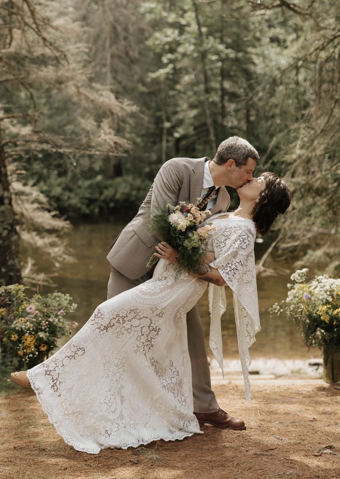 A couple sharing a kiss outdoors by a creek, with trees in the background. The man is leaning over the woman, who is holding a bouquet of flowers and wearing a white lace dress. The scene is surrounded by floral arrangements.