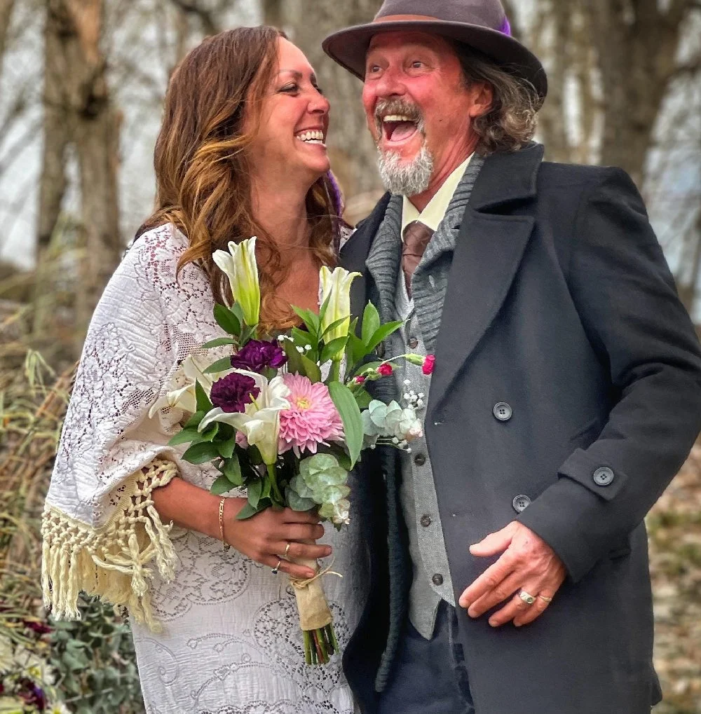 A woman in a white lace dress holding a bouquet of flowers, smiling and looking at a man wearing a black coat and a hat, both outdoors with trees in the background, sharing a joyful moment.