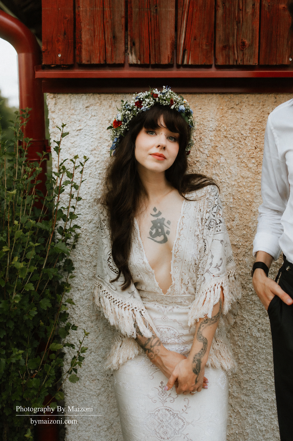 A woman with long dark hair, tattoos, and a floral crown, wearing a crochet white dress, stands against a textured beige wall with greenery to her side.