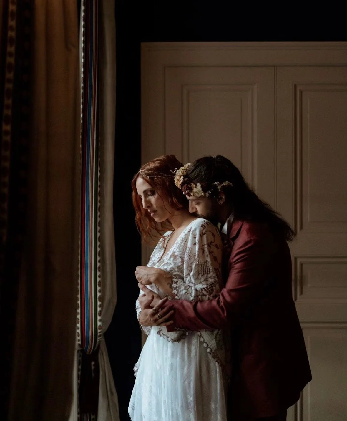 A couple dressed in wedding attire embracing near a window with curtains, one with red hair and a lace dress, the other with long dark hair, floral headpiece, and a maroon suit.