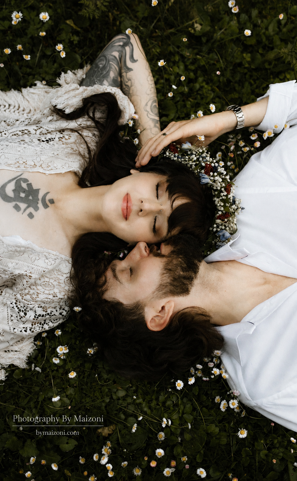 A couple lying on a bed of grass with small white daisies, facing each other with their heads touching. The woman has dark hair, tattoos, and a flower crown, and is wearing a lace dress. The man has curly hair, a beard, and is wearing a white shirt. 