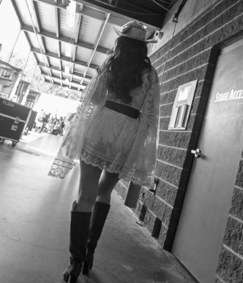 A woman walking in a backstage area, wearing a lace dress, knee-high boots, and a hat, carrying a handbag.