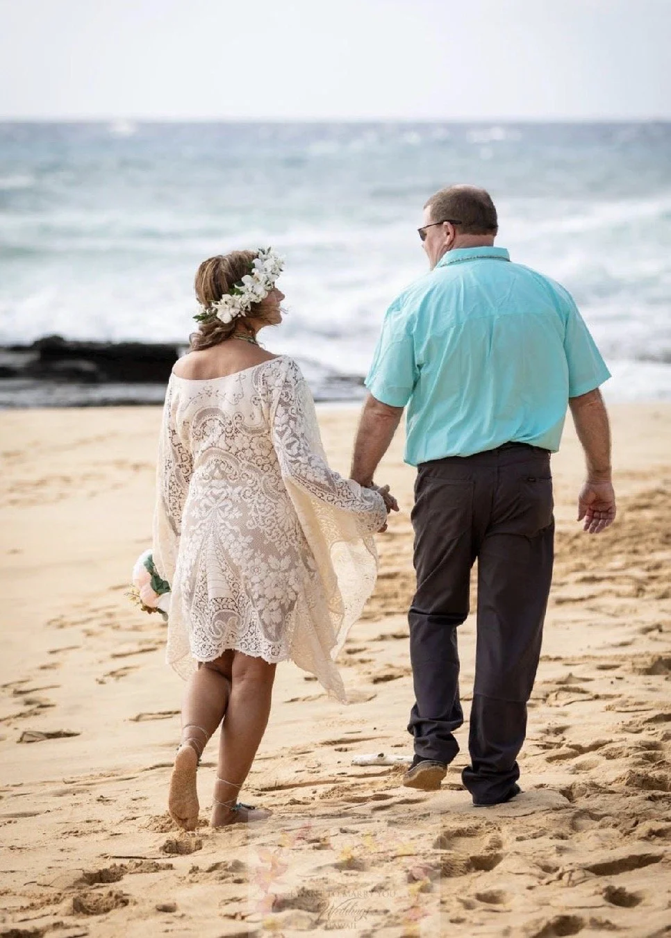 A woman wearing a white lace dress and a floral crown walks hand-in-hand with a man in a blue shirt along a sandy beach, with the ocean in the background, suggesting a wedding or romantic occasion.