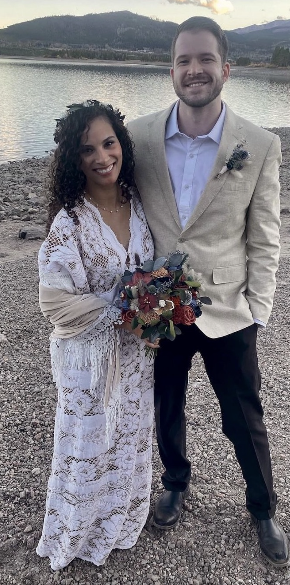 A happy couple on their wedding day standing on a rocky lakeshore with mountains in the background. The woman is wearing a white lace dress and holding a colorful bouquet, while the man is dressed in a light-colored suit with a boutonnière.