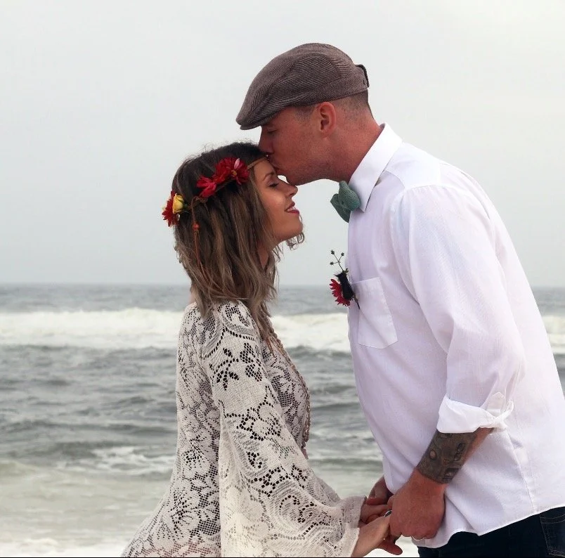 A couple standing on the beach, the woman with a floral headband and lace dress, the man with a flat cap, kissing each other's foreheads, holding hands.