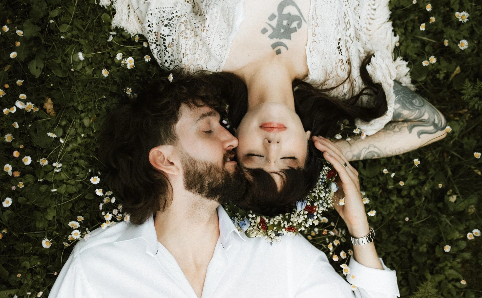 A man and woman lying down on a grassy area with small white daisies, with the woman resting her head on the man's shoulder. The woman has black hair, several facial piercings, and is wearing a floral crown and lace dress. The man has long hair, a beard, and is wearing a white shirt.