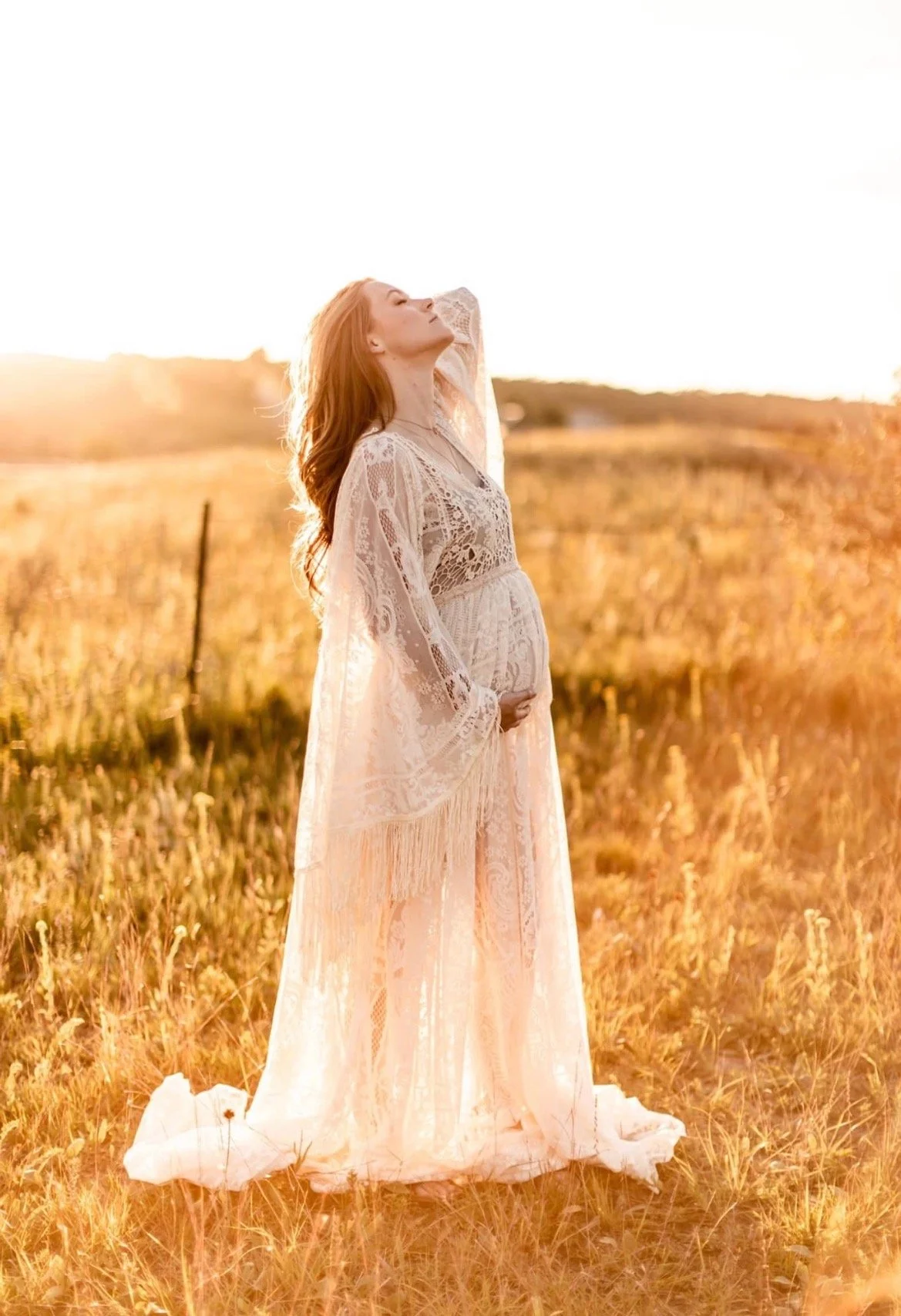 A pregnant woman dressed in a flowing lace gown standing in a golden field at sunset, with her eyes closed and hand on her belly.