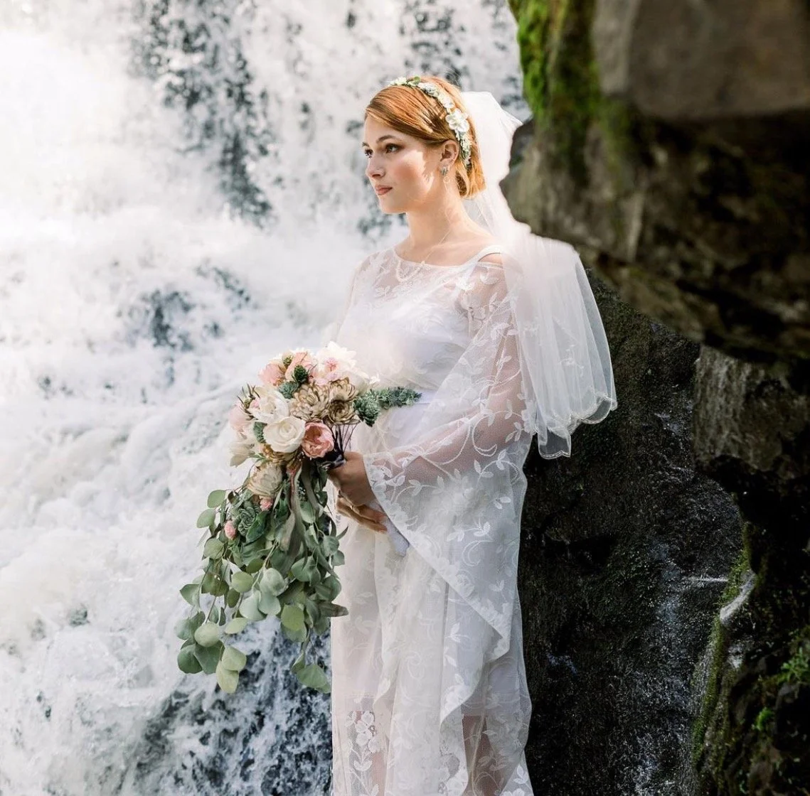 A bride in a lace wedding dress holding a bouquet of flowers, standing beside rocks and a waterfall.