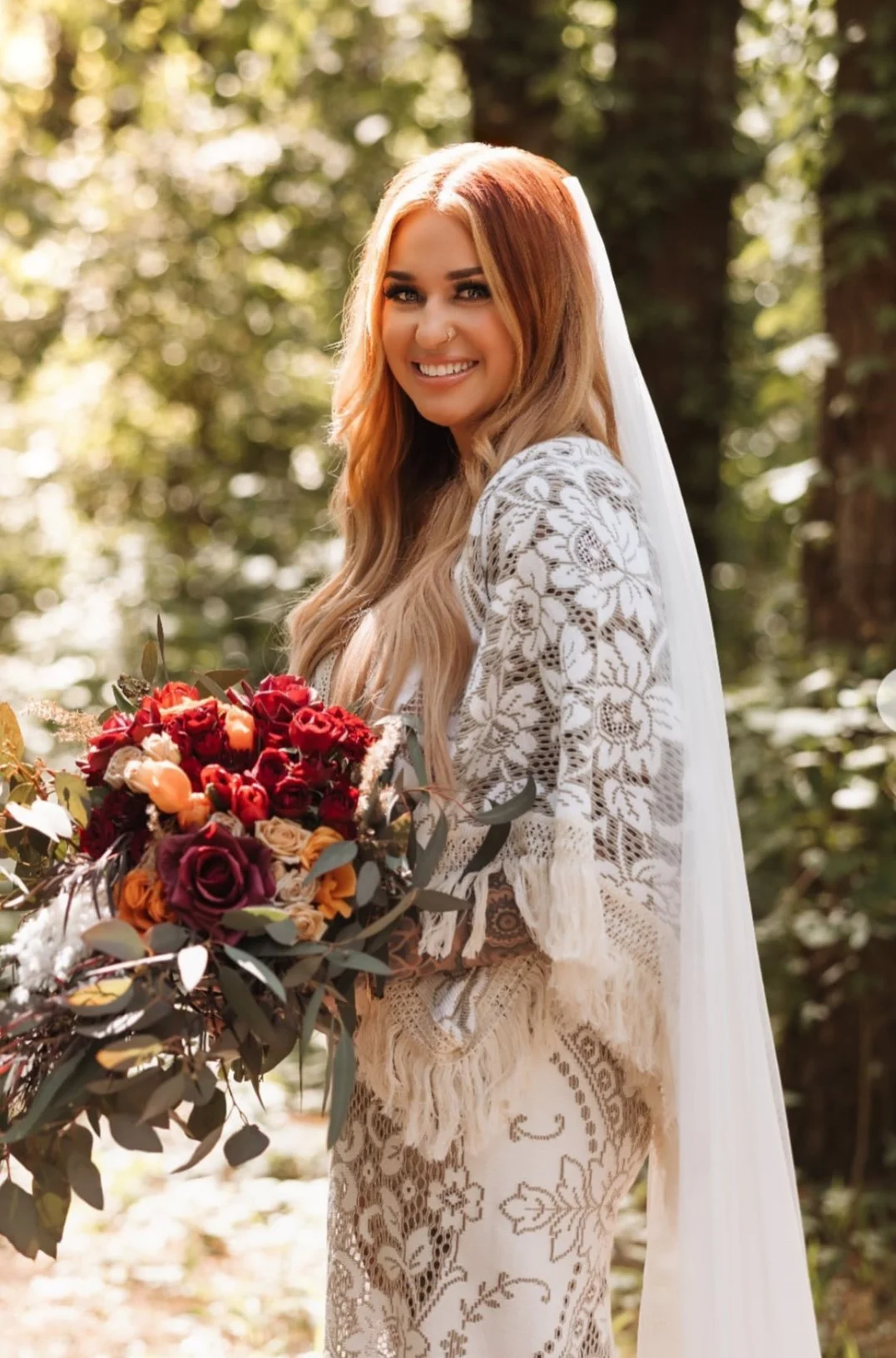 A smiling woman in a wedding dress holding a bouquet of red and peach roses, walking outdoors surrounded by greenery.