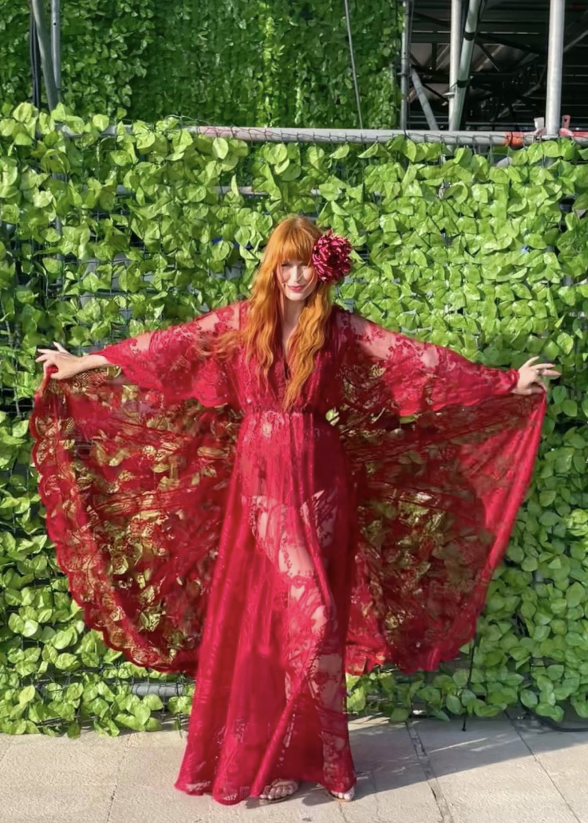 A woman with red hair wearing a flowing red lace dress with wide sleeves, standing in front of a green leafy wall, with a large red flower on her face, smiling and posing with arms outstretched.