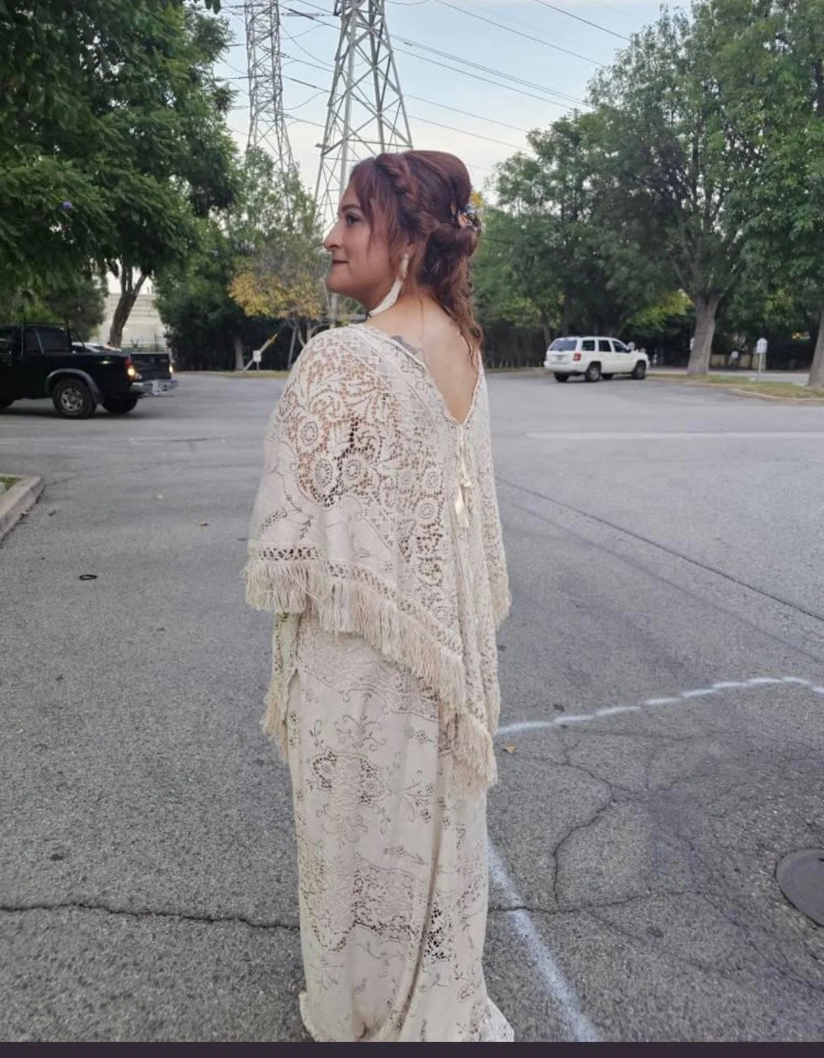 A woman in vintage white lace bohemian attire standing in a parking lot with trees and electrical towers in the background, during daytime.