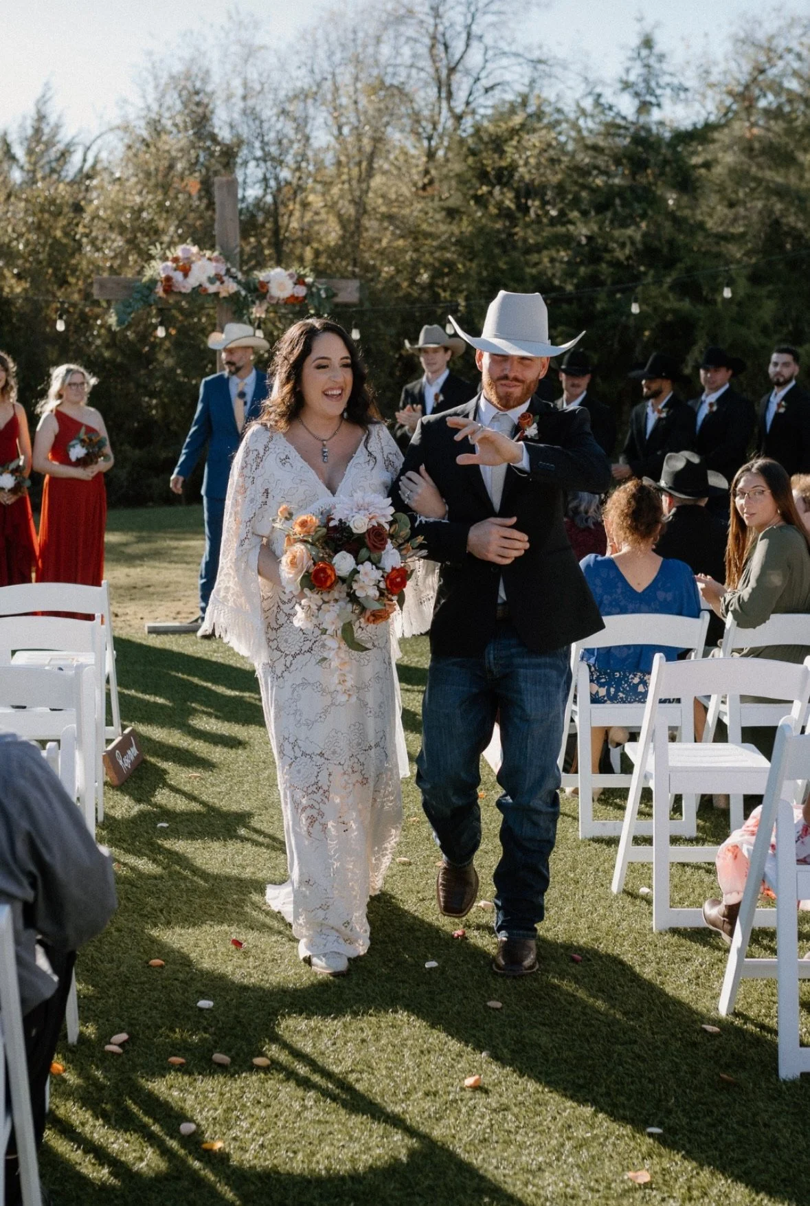 A bride and groom walking down the aisle at an outdoor wedding ceremony. The bride is holding a bouquet and wearing a lace dress, and the groom is wearing a cowboy hat, black blazer, and jeans. Guests are seated on either side, with some standing in the background near a cross decorated with flowers.