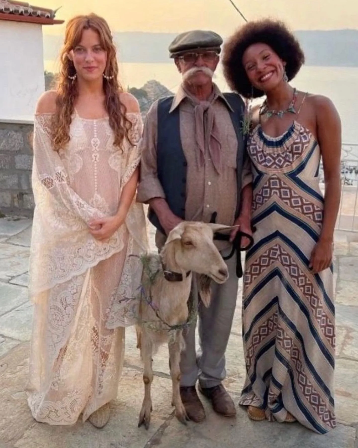 Three women and a man standing outdoors with a goat. The women are wearing colorful dresses, and the man is holding the goat by a leash. The background features a fence, some buildings, and a mountain with a sunset sky.