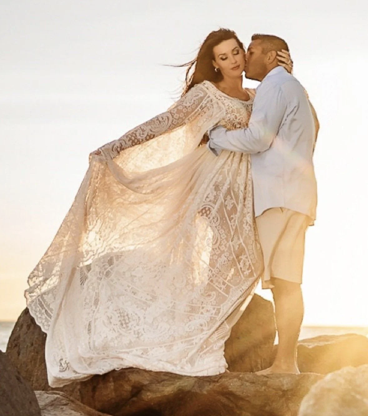 A man and woman embrace on rocks at sunset, the woman wearing a long, flowing white lace dress and the man in a light shirt and shorts.