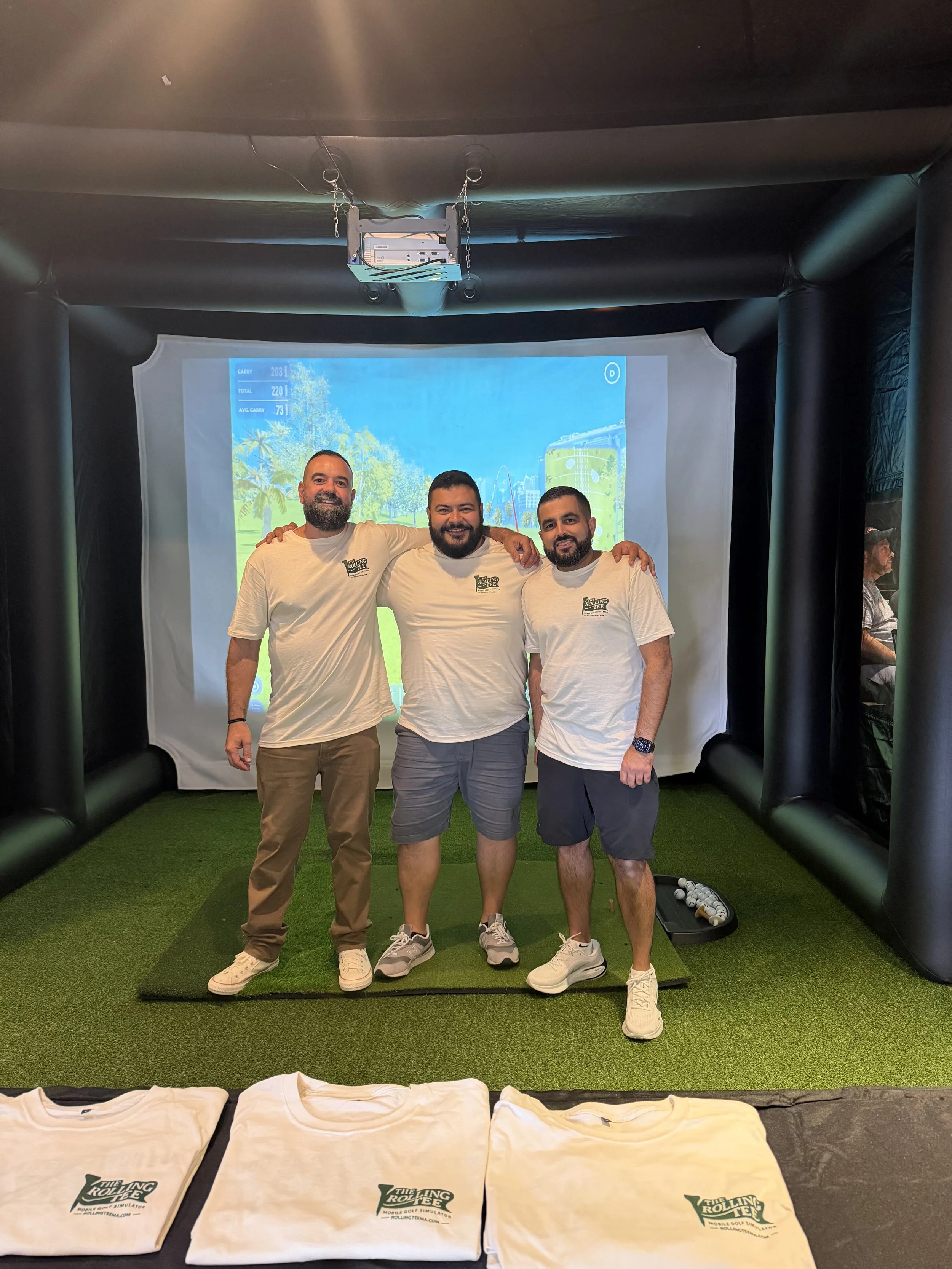 Three men standing inside a mobile golf simulator with a projected screen showing a golf course scene behind them. They are smiling and have their arms around each other's shoulders. In the foreground, there are several rolled-up T-shirts with the logo "The Rolling Tee" on them.