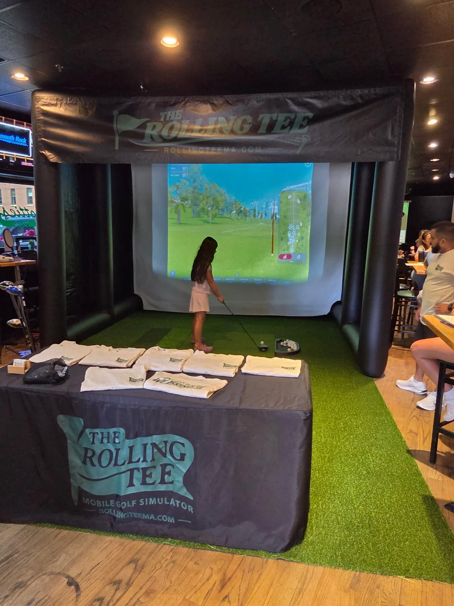 A young girl playing a mobile golf simulator at an indoor venue, with promotional materials from Rolling Tee on a table in the foreground.