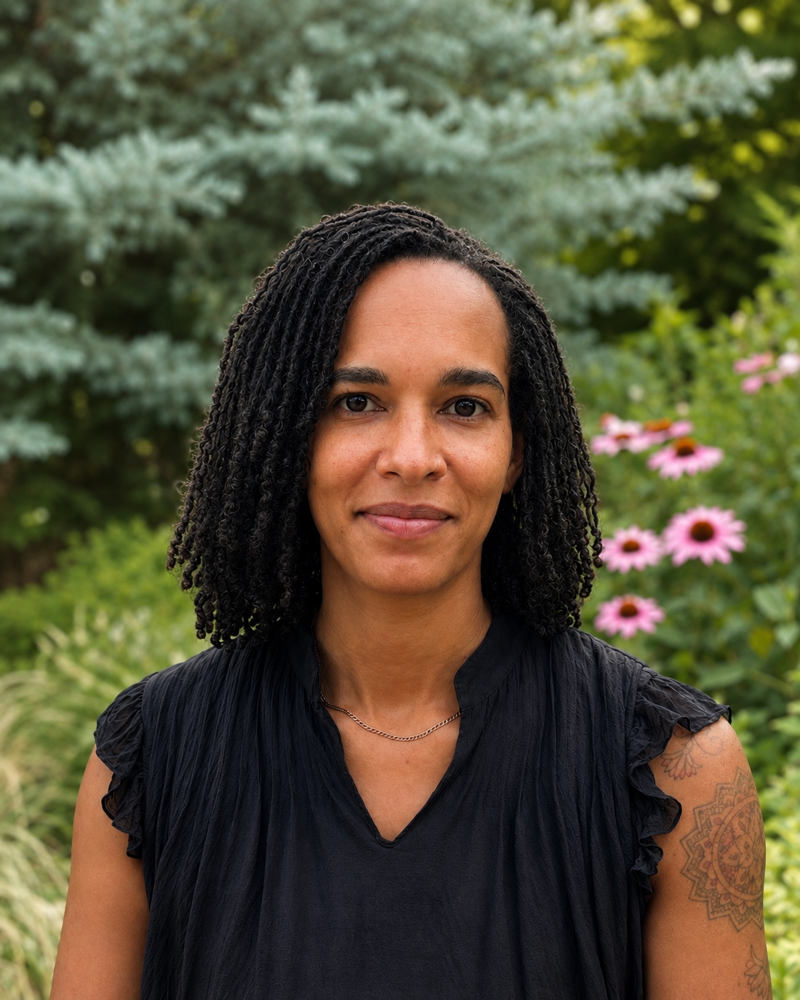 A woman with shoulder-length curly black hair wearing a black top standing outdoors with pink coneflowers flowers and evergreens in the background.